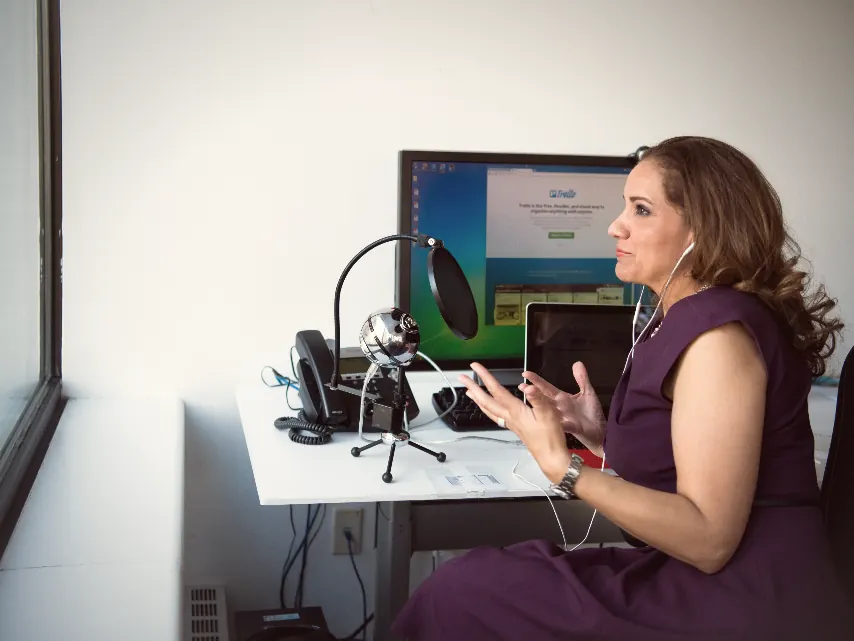 A woman speaking to camera at a desk setup with a microphone